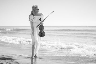 Smiling beautiful woman holding violin while standing on shore at beach