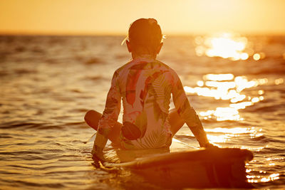Rear view girl sitting on a surfboard during sunset, calm sea without waves of orange light