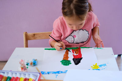 Little girl sits by desk and paints a picture with acrylic paints.