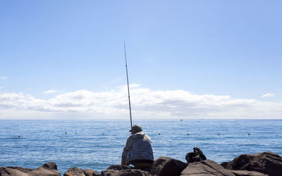 Rear view of man fishing while sitting on rocks by sea against sky