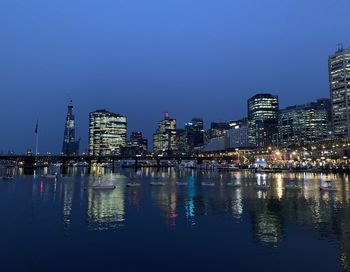 Illuminated buildings by river against clear sky at night