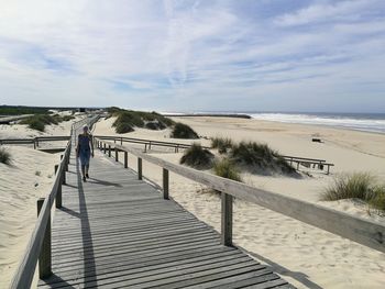 Rear view of woman walking on boardwalk at beach against sky