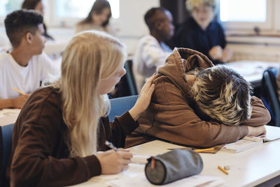 Blond teenage girl consoling depressed female friend resting head on desk in classroom
