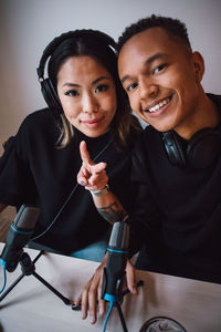 Portrait of smiling female blogger showing peace sign with male colleague sitting against wall at home