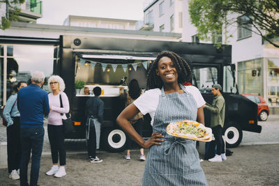 Portrait of smiling female owner with indian food plate standing in city