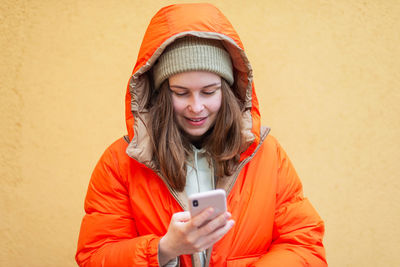 Young woman using phone while standing against orange wall