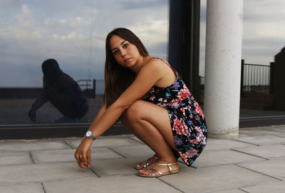 Portrait of young woman crouching on footpath reflecting in mirror outdoors