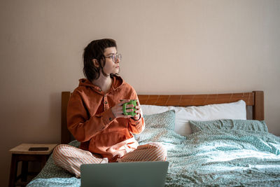 Woman freelancer drinking tea having break sitting in bed at home looking at window.