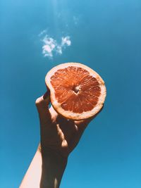 Cropped hand of woman holding grapefruit against blue sky during sunny day