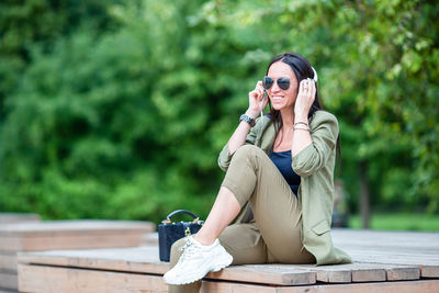 Woman wearing sunglasses and headphones sitting on plank outdoors