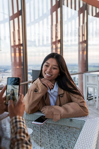 Young woman using mobile phone while sitting on window