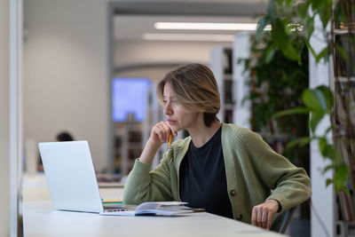 Focused pensive female university teacher preparing for online lecture on laptop in modern library