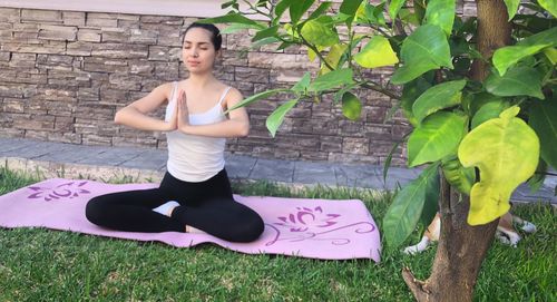 Full length of young woman sitting on green plant against stone wall