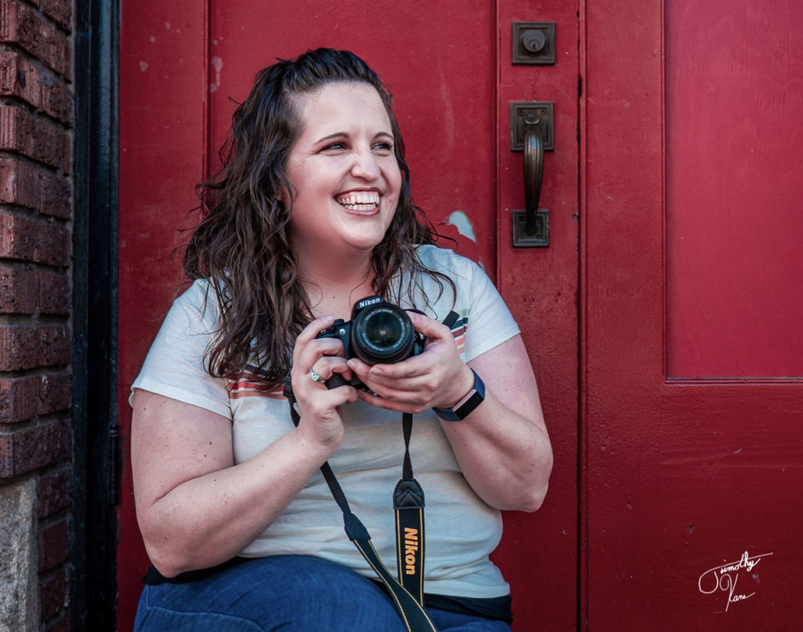 PORTRAIT OF SMILING YOUNG WOMAN HOLDING CAMERA AT ENTRANCE