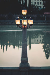 Illuminated street light by lake against sky at sunset