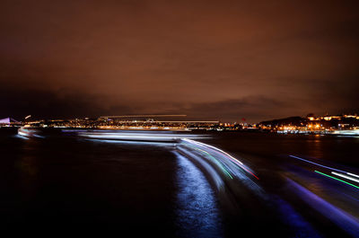 Light trails on road by illuminated city against sky at night
