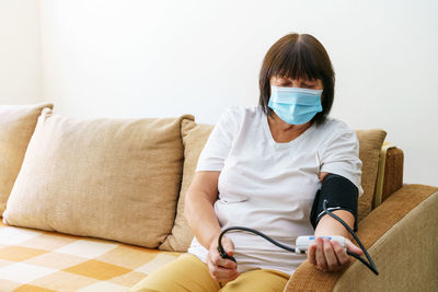 An elderly woman wearing mask measures her blood pressure with an electronic