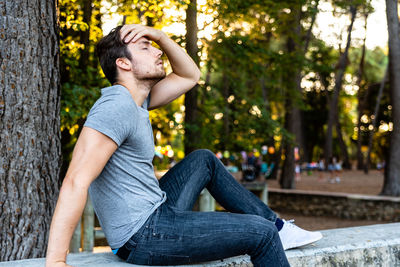 Side view of young man sitting on railing against trees at park