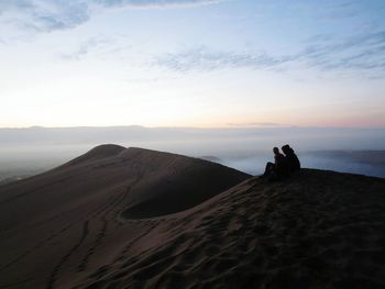 Friends sitting on sand dune against sky during sunset