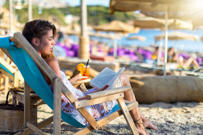 Side view of young woman reading book while sitting on deck chair at beach