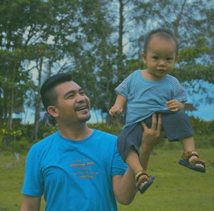 Cheerful father holding son on hand against tree