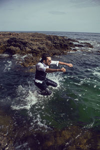 Man with a beard and sunglasses in clothes a vest and a white shirt jump in the sea among the rocks