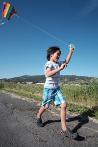 Cheerful boy holding kite running on road by grass against clear sky