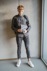 Full length portrait of smiling blond male student wearing eyeglasses holding book leaning on gray wall