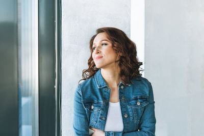 Portrait of smiling charming young woman in jeans jacket near window in modern building