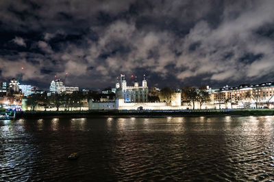 Illuminated buildings by river against sky at night