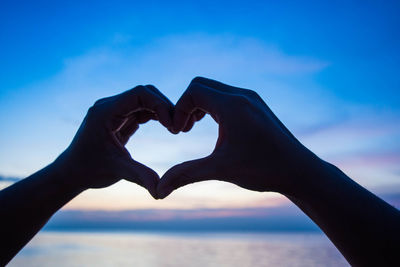 Close-up of hand making heart shape against sky during sunset