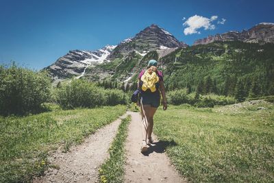 Full length rear view of woman with backpack walking on dirt road against mountains