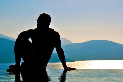 Silhouette of young man sitting in front of lake at sunset