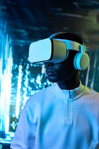 Focused young black man in white t shirt and vr headset watching video in modern room with neon blue lights