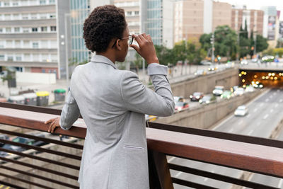 Back view of anonymous african american female executive in classy outfit and eyeglasses standing on balcony and looking at highway and cityscape