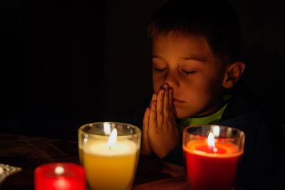 Close-up of boy praying by candles in dark room