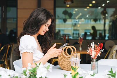 Smiling brunette young woman in white clothes using mobile phone with straw bag at terrace of cafe