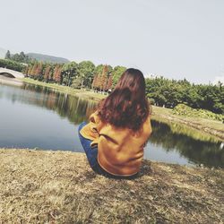 Rear view of woman sitting by lake against clear sky