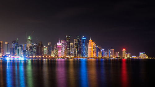 Illuminated city by bay against sky at night