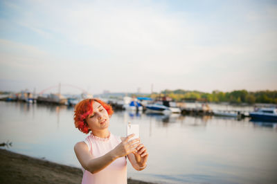 Mid adult woman taking selfie with mobile phone while standing by river against sky