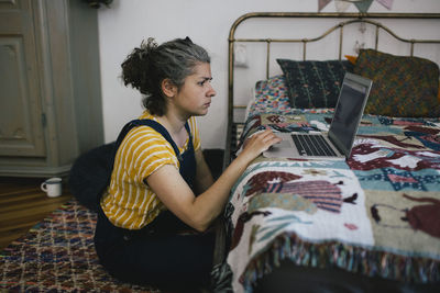 Woman sitting on floor using laptop in bedroom at home