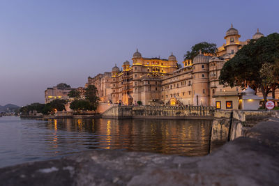 Illuminated building by river against sky in city