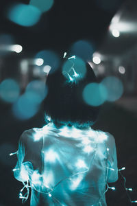 Rear view of woman with illuminated string lights standing in city at night