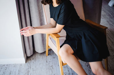 Midsection of young woman in black dress sitting on armchair at home