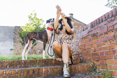 Woman using smart phone while sitting with dog by brick wall