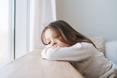 Cute bored thinking little girl with long hair in home clothes near window at home