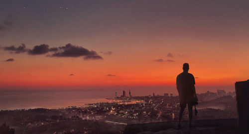 Rear view of silhouette man standing by cityscape against sky during sunset