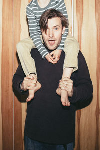 Portrait of father carrying son on shoulder against wooden wall at home