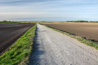 Empty road amidst plowed fields against sky