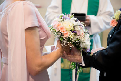 Midsection of bride and bridegroom holding bouquet by priest at church